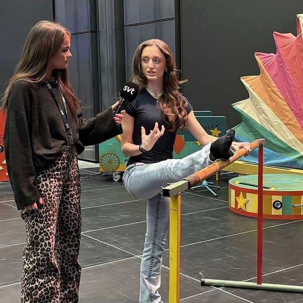 Reportern Lina Dahlberg och Amanda Chilli, elev på Svenska Balettskolan i Göteborg står på en scen där reportern håller en mikrofon, och den eleven gör en balansövning med ett upphöjt ben. Bakgrunden inkluderar färgglada rekvisita och dekorationer, bland annat en stor fläktformad dekoration och lådor med stjärnor.