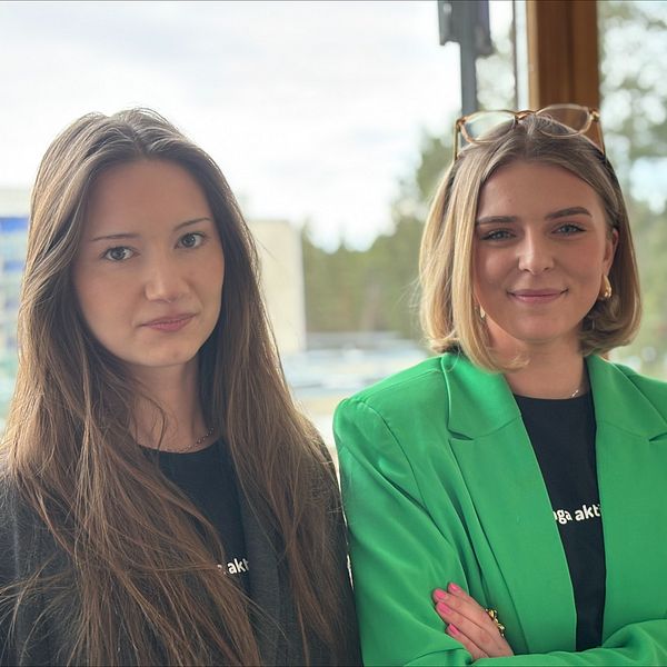 Två personer poserar, en i grön kavaj, mot ett fönster med utsikt över grönskande natur.