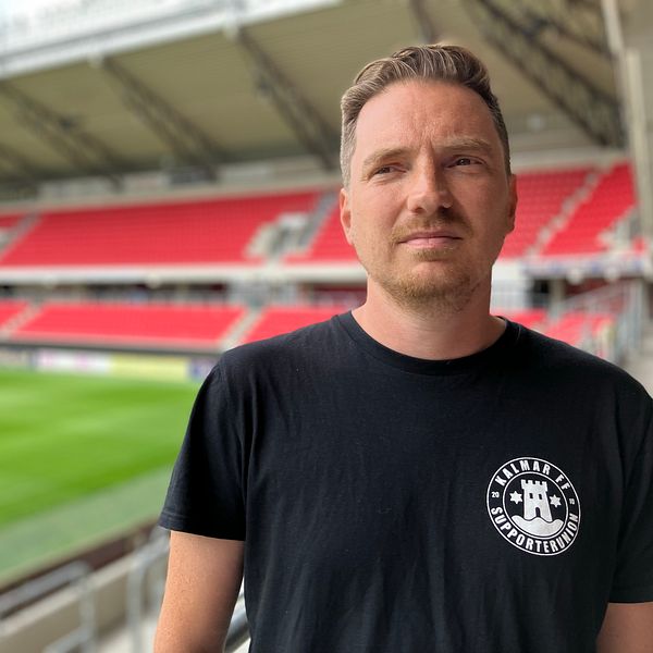 Kortklippt man i svart t-shirt tittar snett till vänster. I bakgrunden syns delar av en fotbollsplan.