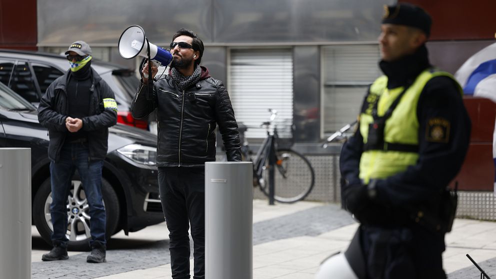 Salwan momika talar utomhus, polisen syns i förgrunden.
