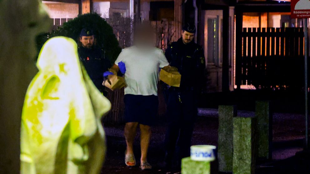 Två poliser eskorterar en man i handfängsel på natten. Ansiktet är suddat av integritetsskäl.