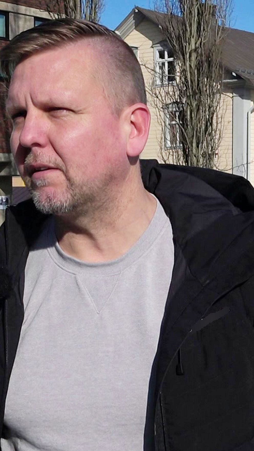 Haparandas kommunalråd Janne Lind står utomhus utanför Haparanda stadshus. Det är soligt och hans svarta jacka är uppknäppt .