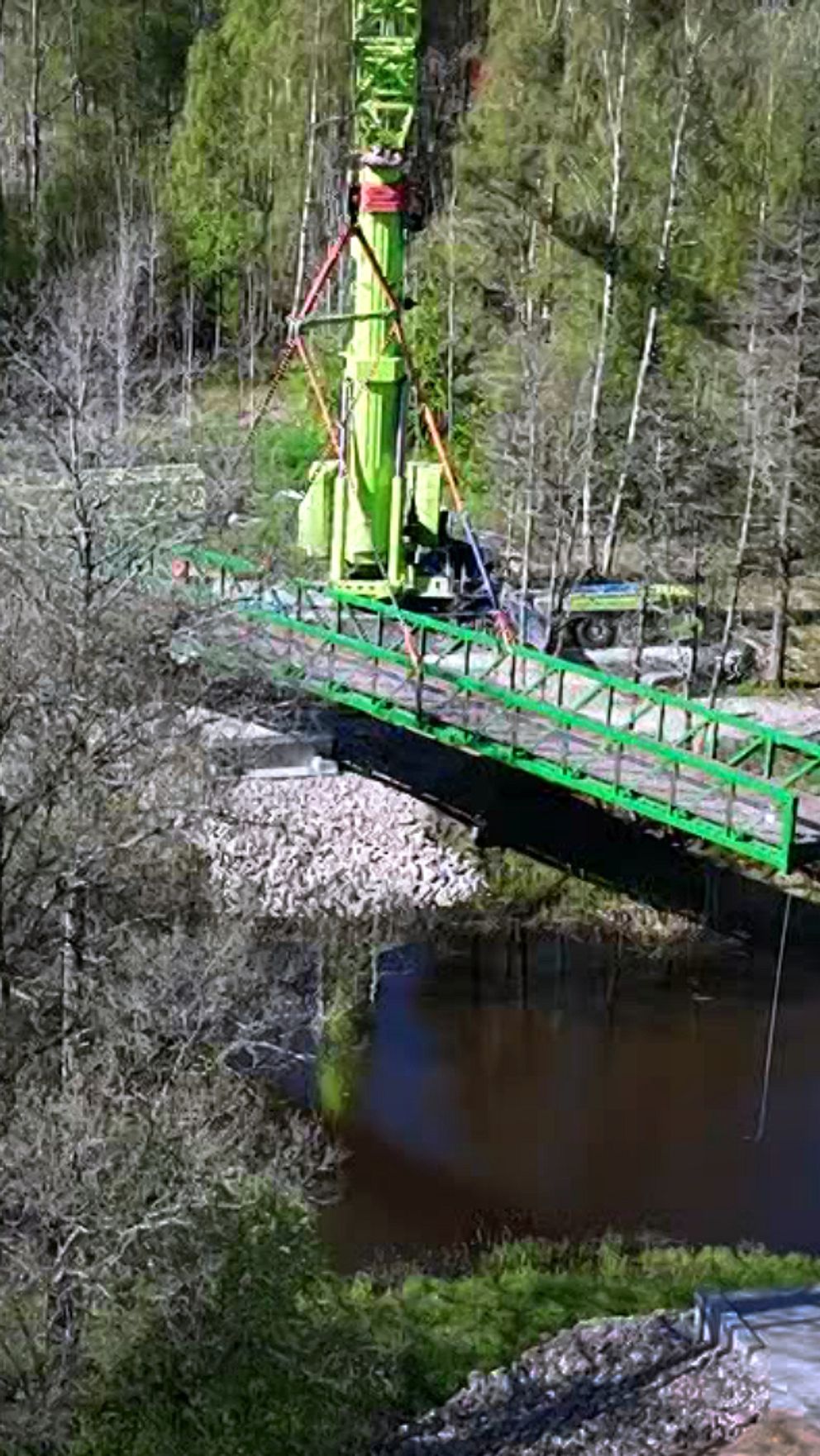 En grön brokran placerar en bro över en skogsklädd å i vårlandskap.
