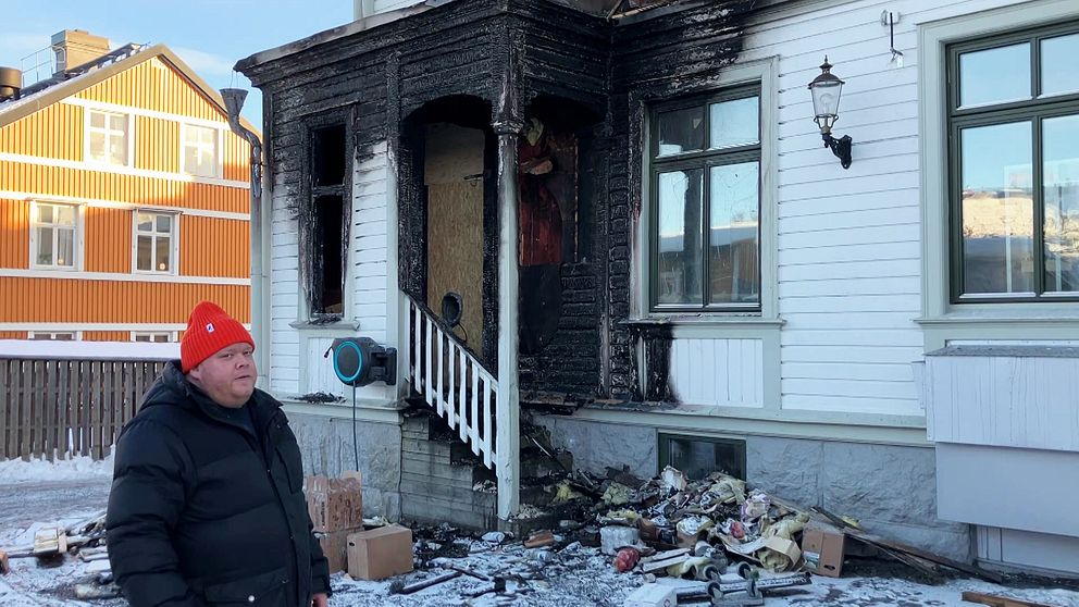 Man i röd mössa står vid brandhärjat hus med sotiga fasaden och bråte på marken.