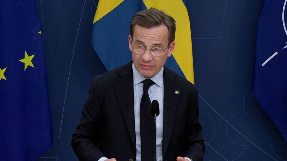 Man vid podium med EU-, svenska och NATO-flaggor i bakgrunden.