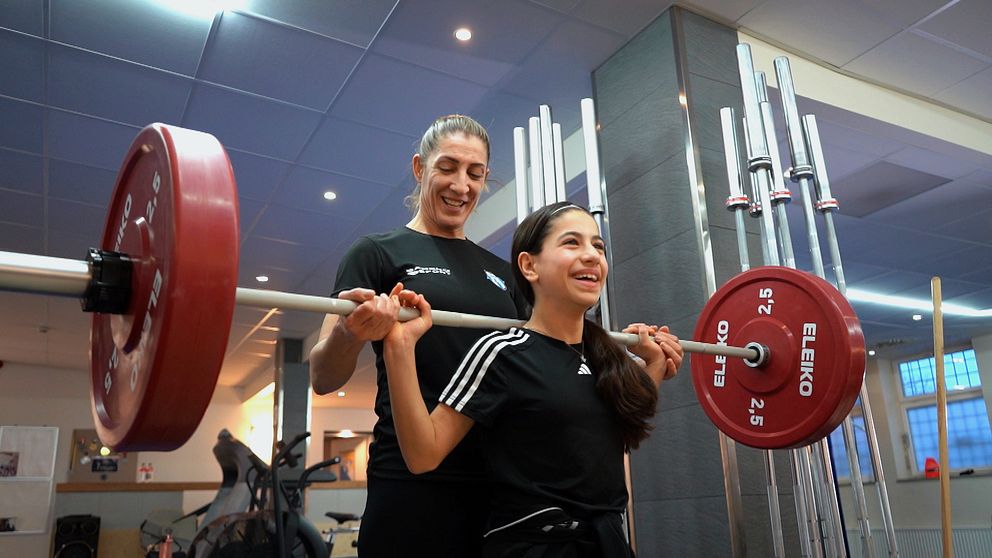 Ung flicka tränar skivstångslyftning med tunga röda vikter i gymmiljö, övervakad av tränaren bakom henne.