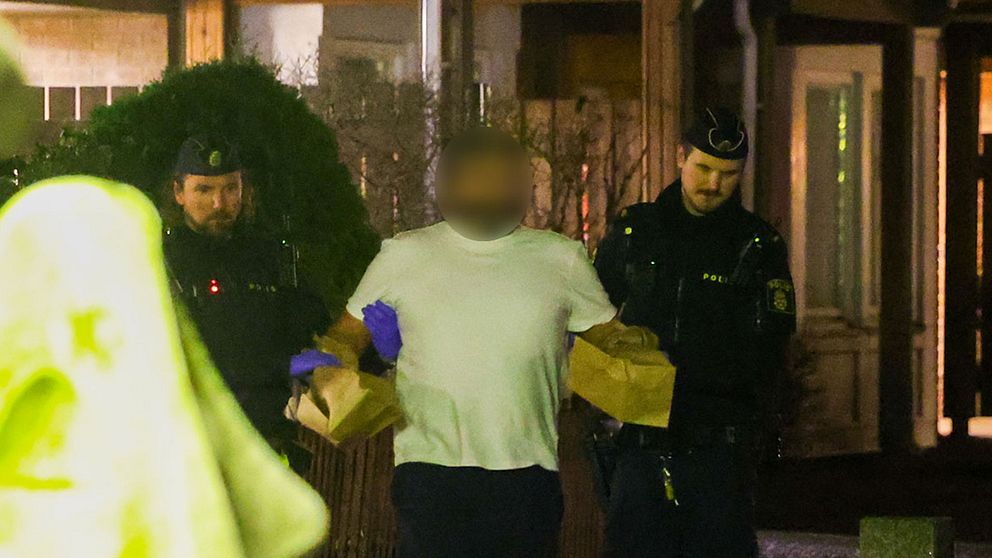 En person vars ansikte har blurrats grips av två poliser. På händerna har personen pappåsar.