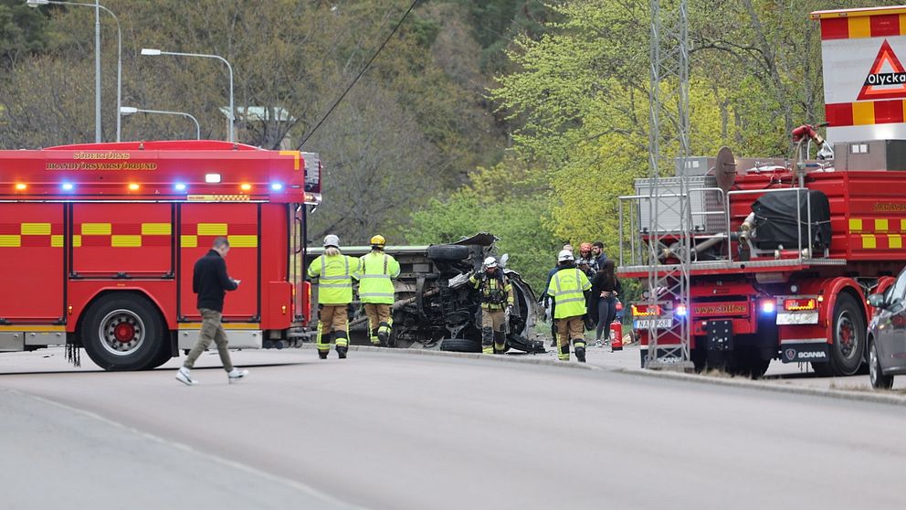 Räddningstjänst arbetar vid en olycksplats med brandbilar och en bil på taket på vägen.
