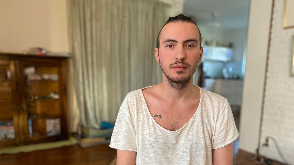 En person sitter på en stol med en ljus, löst sittande t-shirt och mörka byxor. I bakgrunden syns en träig bokhylla och ett rum med köksdetaljer. Golvet är av trä och det finns mjuka ridåer som ramar in rummet.